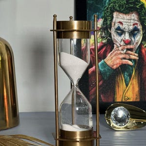 May include: A brass hourglass with white sand. The hourglass is on a grey surface with a gold decorative object and a picture of the Joker in the background.
