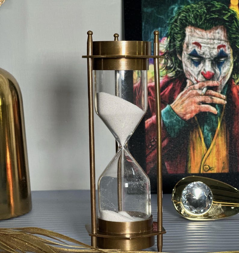 May include: A brass hourglass with white sand. The hourglass is on a grey surface with a gold decorative object and a picture of the Joker in the background.