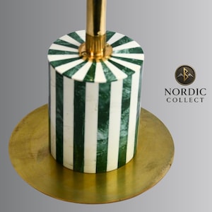 May include: A decorative candle holder with a cylindrical base featuring alternating green and white vertical stripes. The holder has a gold-colored stem and sits on a round, gold-toned base. The logo "NORDIC COLLECT" is visible.