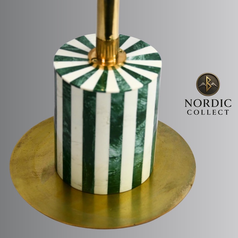 May include: A decorative candle holder with a cylindrical base featuring alternating green and white vertical stripes. The holder has a gold-colored stem and sits on a round, gold-toned base. The logo "NORDIC COLLECT" is visible.