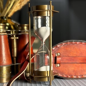 May include: A brass hourglass with a white sand timer. The hourglass is on a table with a leather strap and a pair of binoculars.
