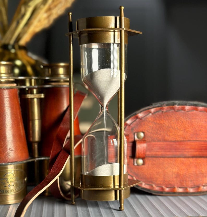 May include: A brass hourglass with a white sand timer. The hourglass is on a table with a leather strap and a pair of binoculars.
