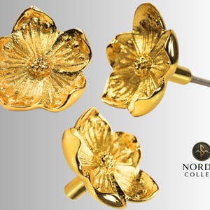 Peut inclure: Trois boutons de meuble dorés en forme de fleur. Chaque bouton présente un motif de pétales détaillé et une zone centrale texturée. Les boutons sont en métal et ont une finition brillante. Le logo de la marque "NORDIC COLLECT" est visible.
