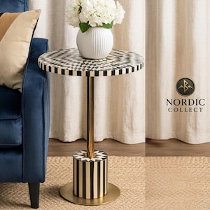Puede incluir: Una mesa auxiliar decorativa con una tapa a cuadros en blanco y negro y una base de metal dorado. La base tiene forma cilíndrica con rayas verticales en blanco y negro. Un jarrón blanco con flores blancas encima. El texto "NORDIC COLLECT" es visible.