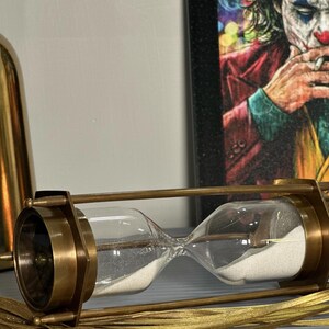 May include: A brass hourglass with a white sand timer. The hourglass is on a gold surface.
