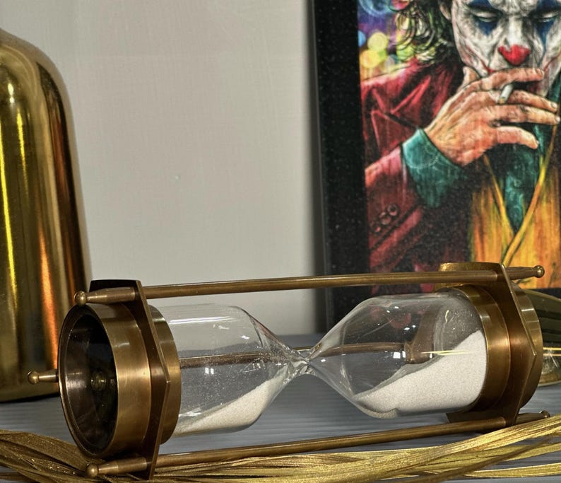 May include: A brass hourglass with a white sand timer. The hourglass is on a gold surface.