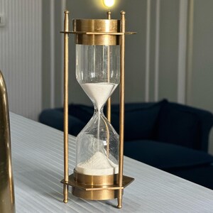 May include: A brass hourglass with a clear glass bulb filled with white sand. The hourglass is on a white surface.