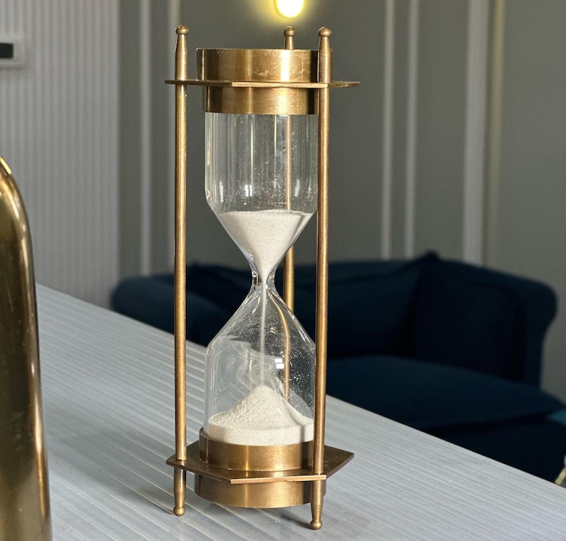 May include: A brass hourglass with a clear glass bulb filled with white sand. The hourglass is on a white surface.