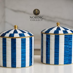 以下が含まれることがあります： 蓋付きの2つのマッチしたセラミックジャー。ジャーは白地に青のストライプと金のトリム。蓋は青と白のチェック柄で、金のノブが付いています。ジャーには「NORDIC COLLECT」のラベルが付いています。