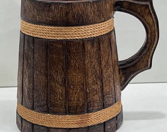 Può includere: Un boccale in legno marrone scuro con un design a botte, caratterizzato da doghe verticali e due fasce orizzontali in corda color marrone chiaro. Il boccale è dotato di un robusto manico in legno marrone scuro fissato sul lato.