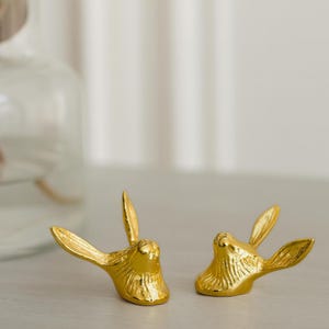 Op de afbeelding: Twee goudkleurige konijnenkopjes met gedetailleerde texturen. De konijnenkoppen kijken in tegengestelde richtingen, met hun oren omhoog. De beeldjes staan op een lichtgekleurd oppervlak, wat wijst op een decoratief of sierlijk doel.