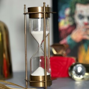 May include: A brass hourglass with white sand. The hourglass is on a table with a red box and a gold object in the background.
