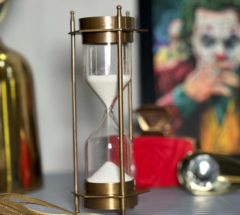 May include: A brass hourglass with white sand. The hourglass is on a table with a red box and a gold object in the background.