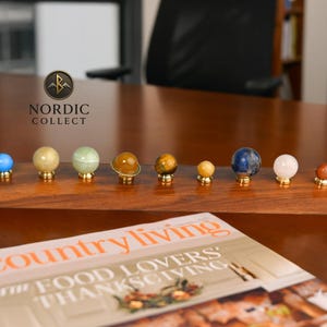 Könnte beinhalten: Eine Holz-Präsentation mit neun Edelstein-Kugeln, jede auf einer goldfarbenen Basis. Die Kugeln sind in verschiedenen Farben gehalten, darunter Blau, Gelb, Grün und Braun. Die Präsentation befindet sich auf einer Holzoberfläche, mit einer Zeitschrift im Vordergrund.