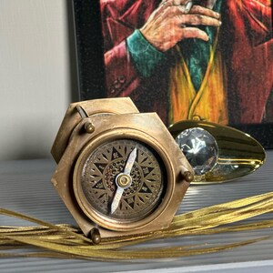 May include: A brass compass with a hexagonal base and a compass rose design. The compass is on a gold surface with a gold and clear crystal object.