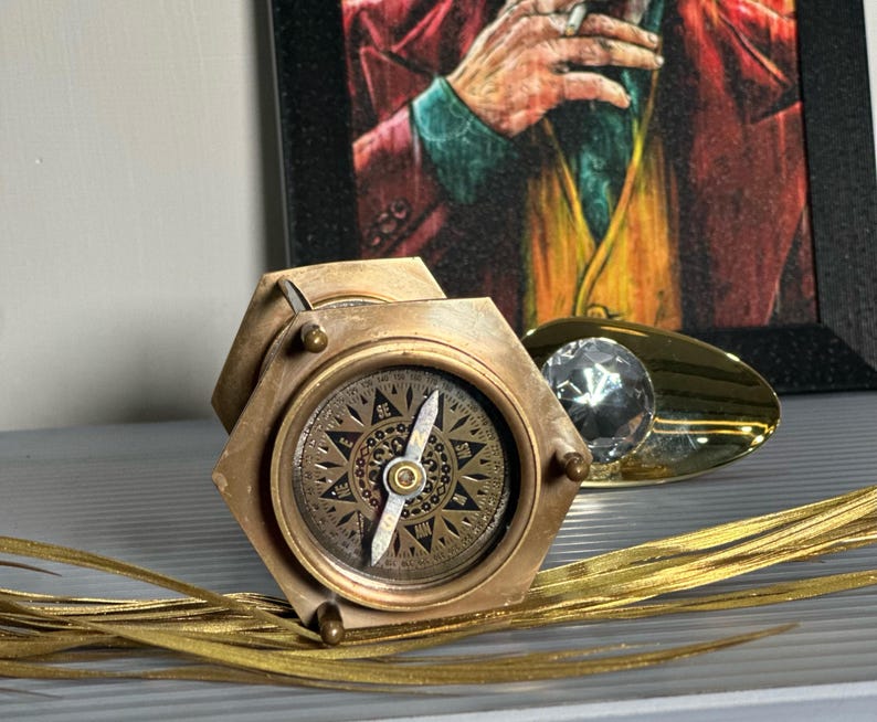 May include: A brass compass with a hexagonal base and a compass rose design. The compass is on a gold surface with a gold and clear crystal object.