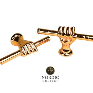 Könnte beinhalten: Zwei goldfarbene Schrankgriffe, jeweils mit einer Hand, die eine horizontale Stange umfasst. Die Griffe haben eine glänzende Oberfläche und sind vor einem weißen Hintergrund platziert. Das Logo "NORDIC COLLECT" ist sichtbar.