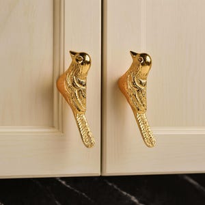 Op de afbeelding: Twee goudkleurige vogelvormige kastgrepen. De handgrepen zijn gemonteerd op een lichtgekleurde kastdeur. De vogelgrepen hebben gedetailleerde veer- en snavelontwerpen en geven de kast een decoratieve uitstraling.