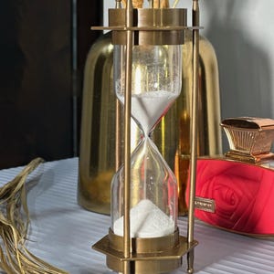 May include: A brass hourglass with white sand, standing on a table with a gold vase and a red bottle with a rose design in the background.