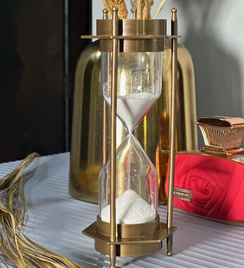May include: A brass hourglass with white sand, standing on a table with a gold vase and a red bottle with a rose design in the background.