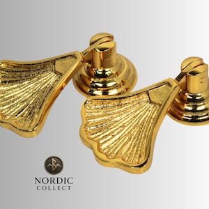 Könnte beinhalten: Zwei goldfarbene Schrankgriffe mit fächerförmigem Design. Die Griffe haben eine strukturierte Oberfläche und sind auf einer runden Basis montiert. Das Logo "NORDIC COLLECT" ist unten sichtbar.