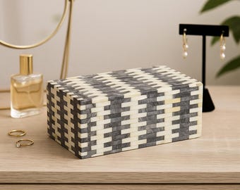 May include: A rectangular decorative box with a geometric pattern in shades of gray and cream. The box is placed on a wooden surface, with a mirror, perfume bottle, and jewelry in the background. The box is a storage item.