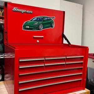 Peut inclure: Un coffre à outils Snap-on rouge vif avec le couvercle ouvert, révélant un autocollant de voiture verte. Le coffre comporte plusieurs tiroirs avec des poignées argentées. Le logo Snap-on est visible sur le couvercle.
