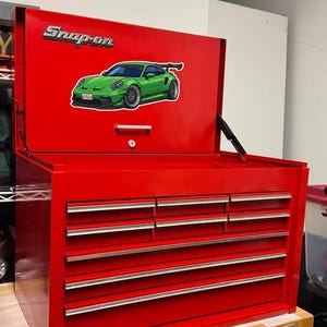 Puede incluir: Un cofre de herramientas Snap-on rojo con la tapa abierta, que muestra una pegatina de un coche deportivo verde. El cofre tiene varios cajones con tiradores plateados. El logotipo de Snap-on se muestra en la tapa.