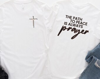 Könnte beinhalten: Weißes T-Shirt mit einem Kreuz auf der Vorderseite und dem Text "The path to peace is always prayer" auf der Rückseite.