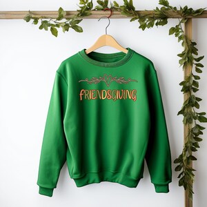 K&ouml;nnte beinhalten: Gr&uuml;ner Pullover mit einer braunen und orangen Grafik, auf der "Friendsgiving" mit einem Herzen und Bl&auml;ttern steht.