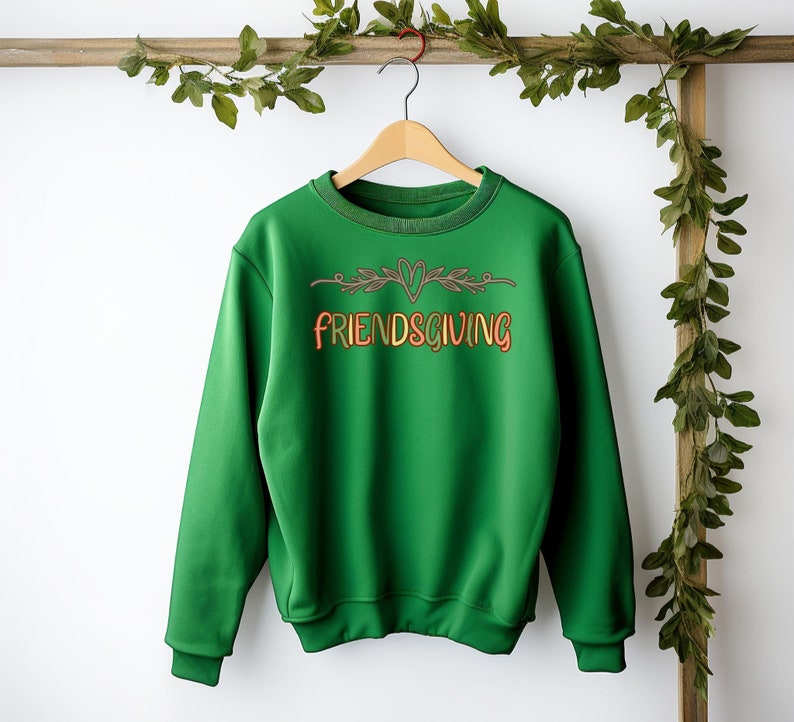 K&ouml;nnte beinhalten: Gr&uuml;ner Pullover mit einer braunen und orangen Grafik, auf der "Friendsgiving" mit einem Herzen und Bl&auml;ttern steht.