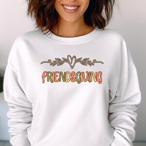 K&ouml;nnte beinhalten: Wei&szlig;er Pullover mit dem Text "FRIENDSGIVING" in bunten Buchstaben mit einem Herz und einem floralen Design &uuml;ber dem Text.