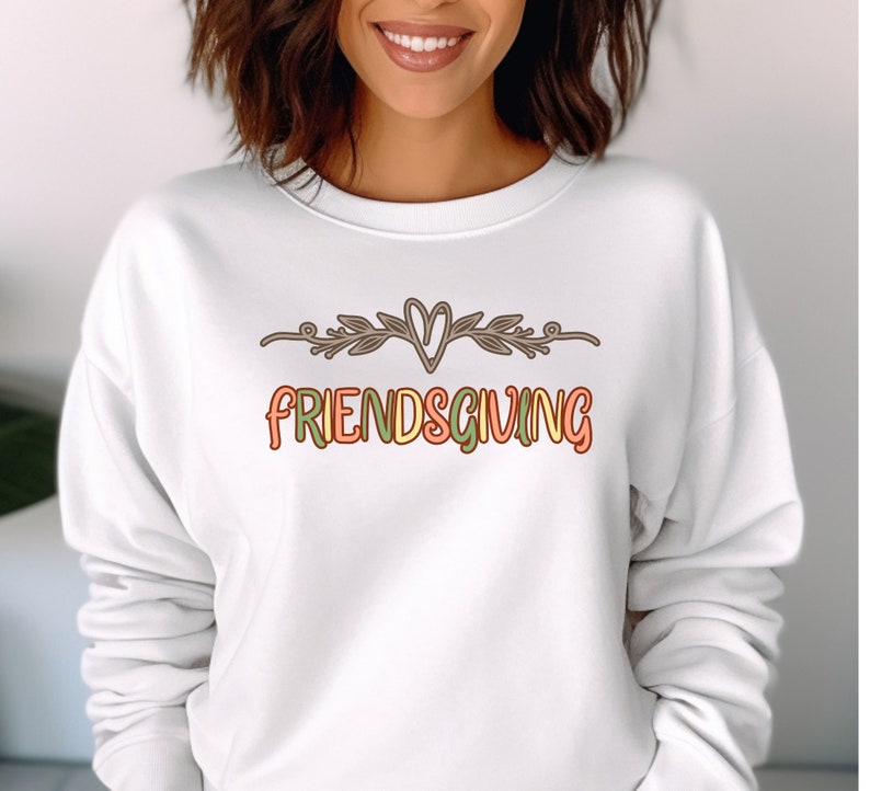 K&ouml;nnte beinhalten: Wei&szlig;er Pullover mit dem Text "FRIENDSGIVING" in bunten Buchstaben mit einem Herz und einem floralen Design &uuml;ber dem Text.