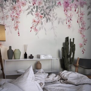 May include: A bedroom interior with a white wall featuring a floral wallpaper with pink blossoms and gray leaves. The room has a bed with white bedding, a wooden chair, a cactus plant, and a shelf with vases and books.