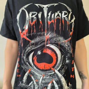 May include: Black t-shirt featuring the band name "Obituary" in white with red dripping accents. The graphic includes a detailed eye design with red and white elements, and the text "A Cause of Death".
