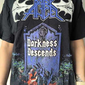 以下が含まれることがあります： 黒いTシャツ。バンド名「Dark Angel」が白と青の文字で、墓石のグラフィックの上に書かれています。墓石には「Darkness Descends」と書かれています。グラフィックには、青と緑の森のシーンが含まれています。