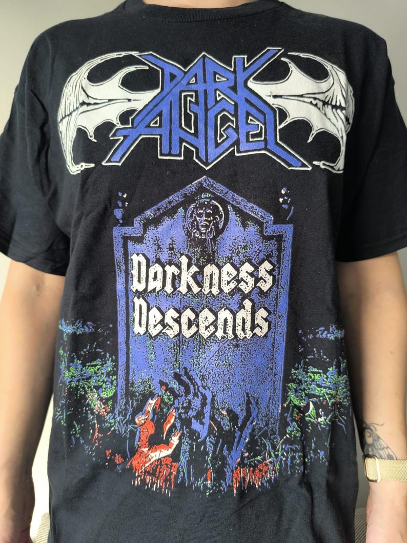 以下が含まれることがあります： 黒いTシャツ。バンド名「Dark Angel」が白と青の文字で、墓石のグラフィックの上に書かれています。墓石には「Darkness Descends」と書かれています。グラフィックには、青と緑の森のシーンが含まれています。
