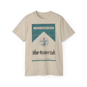 May include: A beige t-shirt with a graphic of a vintage cigarette pack design. The pack is blue and white with the text "Fire of Unknown Origin" and "Blue Öyster Cult". The pack also has a symbol of a cross with a circle around it. The text "8 Class A Tracks" is at the bottom of the pack.