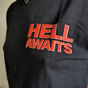 May include: Black t-shirt with the words "HELL AWAITS" in red, outlined in white. The text is centered on the chest. A silver chain necklace is visible at the neckline. The shirt is made of a soft material.