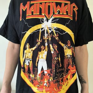 May include: Black t-shirt featuring the band name "Manowar" in orange lettering above a graphic of four figures holding swords. The graphic is set against a yellow circle with lightning bolts and flames.