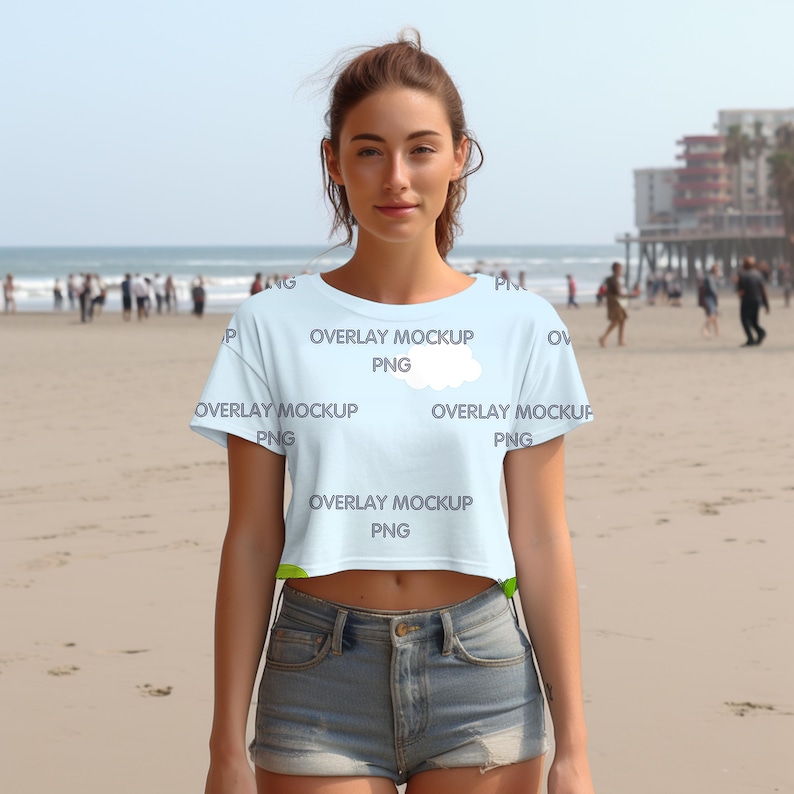 Pu&ograve; includere: Un top corto blu chiaro con una grafica di nuvola bianca e il testo "OVERLAY MOCKUP PNG" ripetuto sul davanti. La modella indossa il top con pantaloncini di jeans e si trova su una spiaggia sabbiosa.