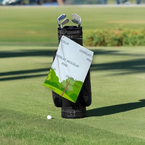 May include: A black golf bag with golf clubs, a white golf ball, and a white towel with green and brown graphics. The towel has the text "OVERLAY MOCKUP PNG". The bag is on a green golf course.