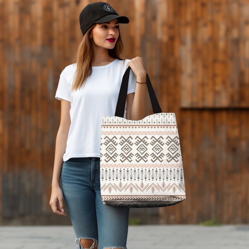 May include: A white tote bag with a brown and pink geometric pattern. The bag has black handles and is being held by a person.
