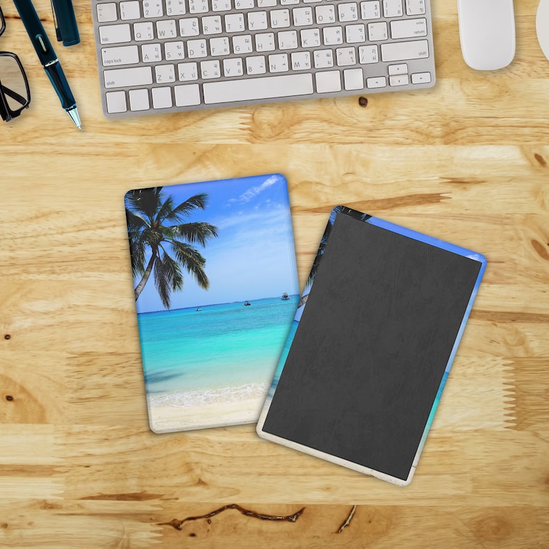 May include: Two rectangular magnets with a tropical beach scene. One magnet shows a palm tree, turquoise water, and white sand. The other magnet has a black surface. The magnets are on a wooden surface.