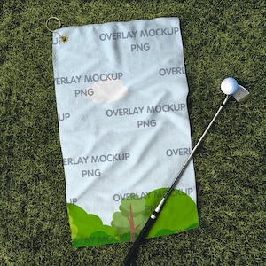 以下が含まれることがあります： 白いゴルフタオルに「Overlay Mockup PNG」という文字が繰り返し書かれています。タオルは緑の芝生の上に置かれ、その隣にはゴルフクラブとボールがあります。