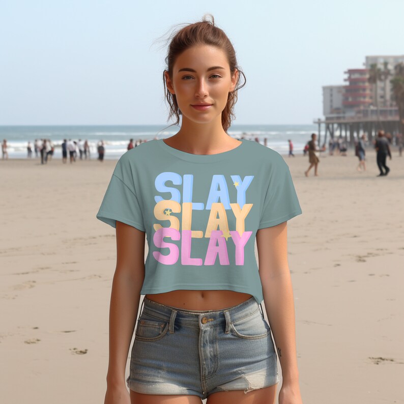 Pu&ograve; includere: Una maglietta corta di colore azzurro chiaro con la parola "SLAY" ripetuta tre volte in diversi colori: blu, giallo e rosa. La maglietta &egrave; indossata da una persona su una spiaggia.