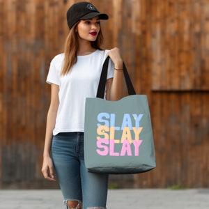 May include: A gray tote bag with black handles and the words "SLAY" repeated three times in different colors: blue, yellow, and pink. The text is stacked vertically.
