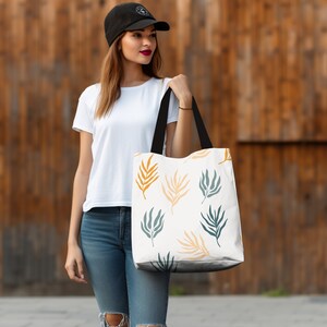 May include: A white tote bag with a pattern of green and yellow leaves. The bag has black handles and is being held by a person wearing a white t-shirt and blue jeans.