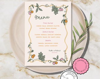 May include: A cream-coloured menu card with floral and fruit illustrations, featuring the word "Menu" at the top. The menu lists courses and dishes, with the names of the couple at the bottom. The card is on a plate with a marble background.