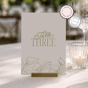 May include: A rectangular table number card in a light beige color featuring the text Table THREE in a dark olive green font. The bottom of the card is decorated with a line art illustration of calla lilies in the same dark olive green color.